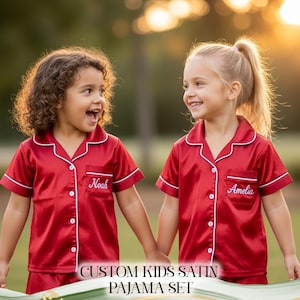 Puede incluir: Dos ni&ntilde;os peque&ntilde;os con conjuntos de pijama de sat&eacute;n rojo a juego con ribetes blancos y nombres personalizados bordados en los bolsillos. El texto "CUSTOM KIDS SATIN PAJAMA SET" se muestra en la parte inferior de la imagen.
