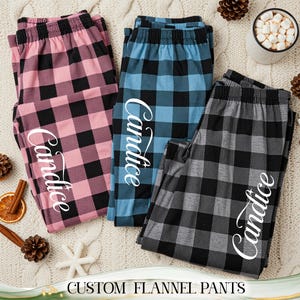 May include: Three pairs of custom flannel trousers in pink, blue, and grey, each with a black plaid pattern and the name "Candice" in white script. The trousers are displayed on a cream-coloured knitted surface, with decorative pine cones and a mug of marshmallows.