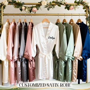 May include: A collection of satin robes in various colors, including ivory, pink, gray, and navy blue, hang on a gold rack. The robes are displayed with floral accents and the text "CUSTOMIZED SATIN ROBE" at the bottom. One robe is personalized with the name "Emliya".