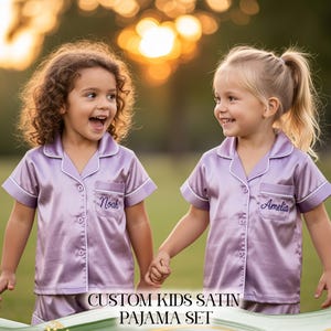 Puede incluir: Dos ni&ntilde;os peque&ntilde;os con conjuntos de pijama de sat&eacute;n lavanda a juego con ribete blanco y nombres personalizados bordados en el bolsillo. Los conjuntos de pijama incluyen una camiseta de manga corta y pantalones cortos. El texto "CUSTOM KIDS SATIN PAJAMA SET" est&aacute; en la parte inferior de la imagen.