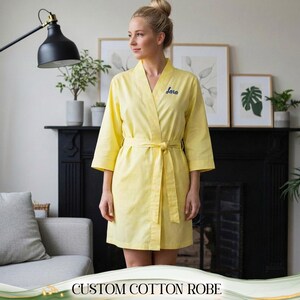 Peut inclure: Peignoir en coton jaune avec une ceinture assortie. Le peignoir a des manches trois-quarts et le nom "Sara" brodé en bleu sur la poitrine. Le texte "CUSTOM COTTON ROBE" est au bas de l'image.