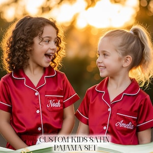 Puede incluir: Dos ni&ntilde;os con conjuntos de pijama de sat&eacute;n rojo con ribetes blancos y bordados personalizados. Un conjunto de pijama est&aacute; bordado con "Noah" y el otro con "Amelia". La imagen incluye el texto "CUSTOM KIDS SATIN PAJAMA SET".