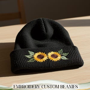 May include: Black knitted beanie hat with an embroidered design of two yellow sunflowers and green leaves. The text "EMBROIDERY CUSTOM BEANIES" is at the bottom of the image.