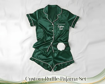 May include: A dark green satin pajama set with white trim, consisting of a short-sleeved button-up shirt and shorts. The shirt has a pocket with the text "Custom Text" and a white flower. The set is displayed on a white surface.