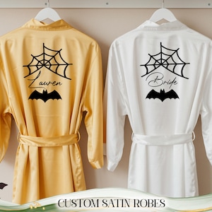May include: Two satin robes, one gold and one white, hang on a rack. The gold robe has a black spiderweb, bat, and the name "Lauren" printed on the back. The white robe has "Bride" printed on the back, with a spiderweb and bat design. The text "CUSTOM SATIN ROBES" is at the bottom.