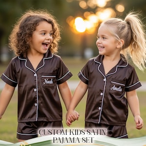 Puede incluir: Dos ni&ntilde;os con conjuntos de pijama de sat&eacute;n marr&oacute;n a juego con ribetes blancos y nombres personalizados. Los conjuntos de pijama incluyen una camisa con botones y pantalones cortos. El texto "CUSTOM KIDS SATIN PAJAMA SET" est&aacute; en la parte inferior de la imagen.