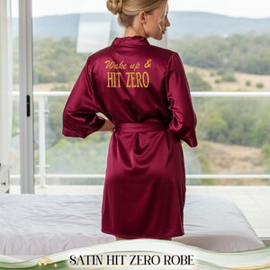 May include: Burgundy satin robe with the text "Wake up & Hit Zero" in gold lettering on the back. The robe has a matching belt and is knee-length. The image shows the robe on a person standing near a window.