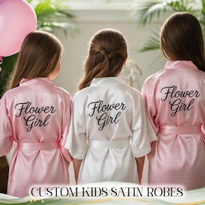 Könnte beinhalten: Drei junge Mädchen tragen Satin-Roben, zwei in Rosa und eine in Weiß. Auf den Roben steht in schwarzer Schrift "Flower Girl" auf dem Rücken. Das Bild zeigt auch rosa und lila Luftballons.