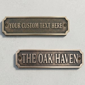 May include: Two bronze-colored rectangular plaques with rounded corners. The top plaque reads "YOUR CUSTOM TEXT HERE" in raised lettering. The bottom plaque reads "THE OAK HAVEN". Both are mounted with screws.