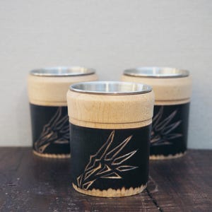 Può includere: Tre bicchieri cilindrici con parte superiore in legno chiaro e base nera. La base nera presenta un disegno intagliato. I bicchieri hanno un interno color argento e sono appoggiati su una superficie di legno scuro.