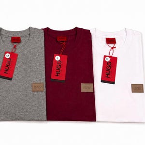 May include: Three crew neck t-shirts in heather grey, dark red, and white. Each shirt features a small rectangular brown patch on the lower left side with the text HUGO. Red paper tags are attached to the collars with the text HUGO and size labels M, L, XL.