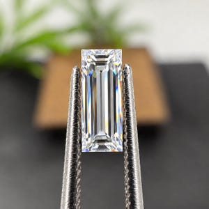 May include: A clear, rectangular baguette-cut gemstone with sharp, linear facets that create a bright, reflective surface. The stone is held vertically by a pair of textured, silver-toned metal tweezers, showcasing its high clarity and brilliance.