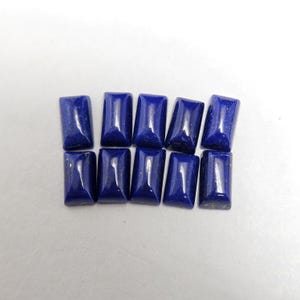 May include: Ten rectangular, polished lapis lazuli gemstones are arranged on a light surface. The stones are a deep, rich blue with subtle golden flecks, and have a smooth, reflective surface. The gemstones are of similar size and shape.