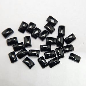 Peut inclure: Plusieurs pierres précieuses noires, rectangulaires et brillantes, sont dispersées sur une surface blanche. Les pierres ont des bords arrondis et reflètent la lumière, suggérant une finition polie. Ces pierres sont pour la joaillerie.