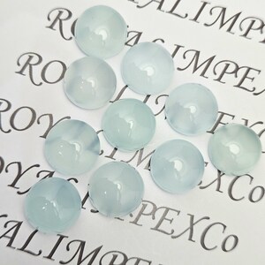 K&ouml;nnte beinhalten: Eine Ansammlung von zehn runden, hellblauen Cabochon-Edelsteinen. Die polierten Steine sind durchscheinend und auf einer wei&szlig;en Oberfl&auml;che mit dem Aufdruck "ROYAL IMPEX Co" in dekorativer Schrift angeordnet.