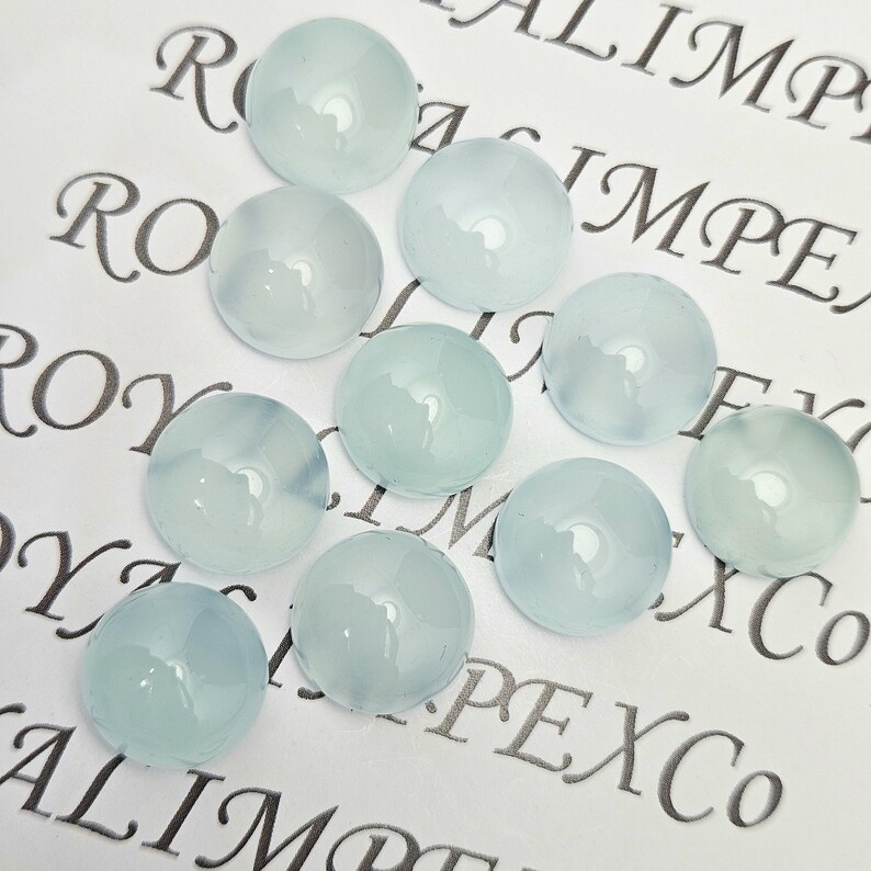 K&ouml;nnte beinhalten: Eine Ansammlung von zehn runden, hellblauen Cabochon-Edelsteinen. Die polierten Steine sind durchscheinend und auf einer wei&szlig;en Oberfl&auml;che mit dem Aufdruck "ROYAL IMPEX Co" in dekorativer Schrift angeordnet.