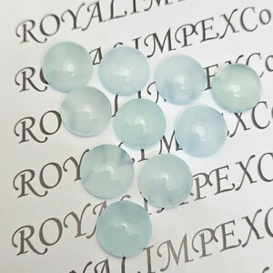 K&ouml;nnte beinhalten: Eine Ansammlung von neun runden, hellblauen Cabochon-Edelsteinen. Die Edelsteine sind auf einer wei&szlig;en Oberfl&auml;che angeordnet, auf der der Text "ROYAL IMPEX CO" in Schwarz gedruckt ist. Die Edelsteine haben eine glatte, polierte Oberfl&auml;che.