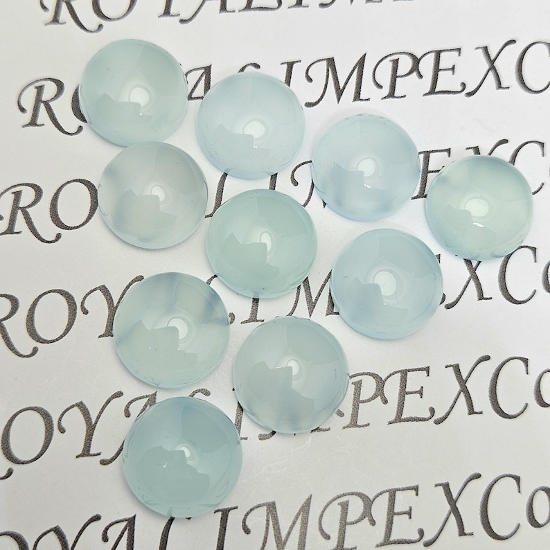 K&ouml;nnte beinhalten: Eine Ansammlung von zehn runden, hellblauen Cabochons. Die Cabochons sind auf einer wei&szlig;en Oberfl&auml;che mit aufgedrucktem Text angeordnet. Die Edelsteine haben ein glattes, poliertes Aussehen und sind leicht durchscheinend.