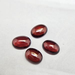 May include: Four polished, oval-shaped, dark red gemstones. The stones have a smooth, reflective surface, and are arranged against a white background. The gemstones are likely for jewelry making or collecting.
