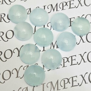 K&ouml;nnte beinhalten: Eine Sammlung von zehn runden, hellblauen Cabochon-Edelsteinen. Die polierten Steine haben eine glatte, gl&auml;nzende Oberfl&auml;che und sind auf einem wei&szlig;en Hintergrund mit gedrucktem Text angeordnet. Die Edelsteine haben eine helle, durchscheinende Farbe.