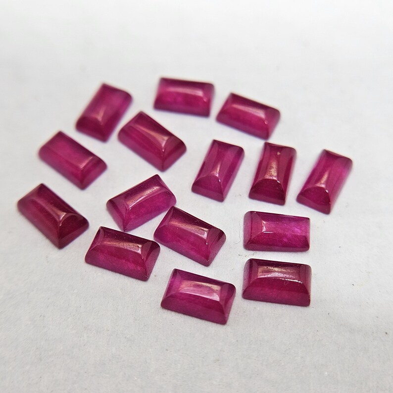 Pode incluir: Uma cole&ccedil;&atilde;o de pedras preciosas de rubi retangulares e polidas. Cada gema tem uma cor magenta profunda e uma superf&iacute;cie lisa e refletora. As gemas est&atilde;o dispostas sobre uma superf&iacute;cie de cor clara, mostrando sua tonalidade e forma vibrantes.