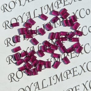 May include: A collection of small, rectangular, deep magenta gemstones scattered on a white surface. The gemstones are translucent and appear to be cut with a polished finish. The background features the text "ROYALIMPEX Co" in a repeating pattern.