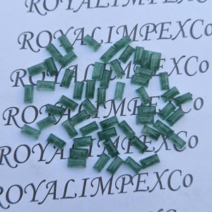 Könnte beinhalten: Eine Ansammlung kleiner, rechteckiger, smaragdgrüner Edelsteine, die auf einer weißen Oberfläche verstreut sind. Die Edelsteine sind durchscheinend und scheinen präzise geschnitten zu sein. Der Hintergrund zeigt den Text "ROYALIMPEXCO".