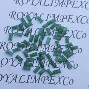 May include: Numerous small, rectangular, emerald-green gemstones are scattered on a white surface. The gemstones are translucent and polished. The background features the text "ROYALIMPEXCo" in a repeating pattern.