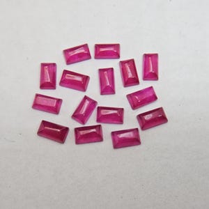 Pode incluir: Uma cole&ccedil;&atilde;o de pedras preciosas de rubi retangulares e facetadas. As gemas s&atilde;o de uma cor vermelho-rosada profunda e vibrante e est&atilde;o espalhadas sobre uma superf&iacute;cie branca. As facetas capturam a luz, criando reflexos e sombras.