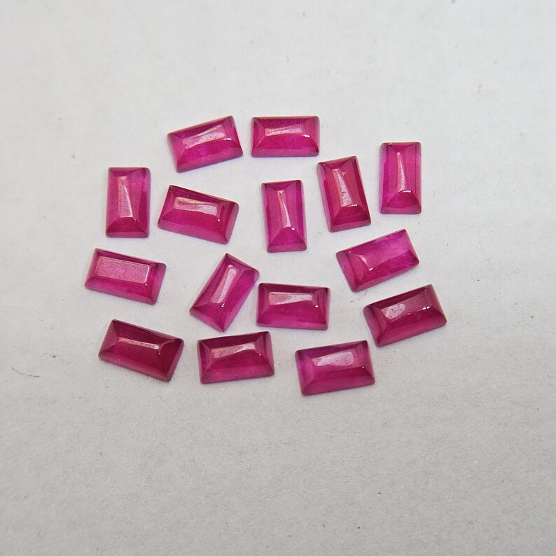 Pode incluir: Uma cole&ccedil;&atilde;o de pedras preciosas de rubi retangulares e facetadas. As gemas s&atilde;o de uma cor vermelho-rosada profunda e vibrante e est&atilde;o espalhadas sobre uma superf&iacute;cie branca. As facetas capturam a luz, criando reflexos e sombras.