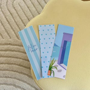 Peut inclure: Trois marque-pages bleus et blancs. L'un présente des rayures verticales et les mots "Bleu mer". Un autre a un motif de petits cœurs bleus. Le troisième représente une scène côtière avec une porte, l'océan et une plante en pot.