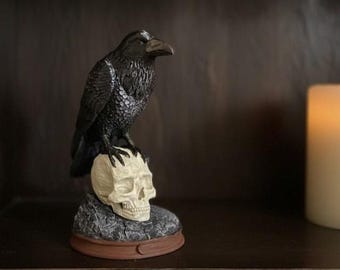 May include: A black raven statue perched atop a cream-colored skull. The raven has detailed feather textures and a prominent beak. The skull rests on a textured gray base with a brown rim, creating a gothic or Halloween theme.