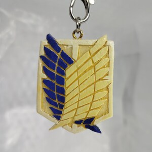May include: A cream-colored keychain pendant featuring the emblem of the Survey Corps from the anime series Attack on Titan. The emblem has blue and yellow wings, with a silver-colored clasp.