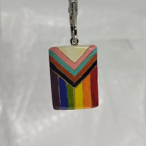 Può includere: Un ciondolo rettangolare con un design a bandiera arcobaleno, con un fermaglio color argento. Il design della bandiera include strisce rosse, arancioni, gialle, verdi, blu, nere, rosa e bianche. Il ciondolo è probabilmente in metallo o materiale simile.