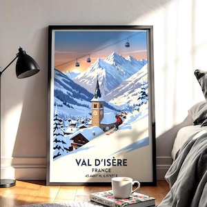May include: A framed art print of Val d'Isère, France, featuring a snowy mountain landscape with a church and skiers. The print has a black frame and includes the text "VAL D'ISÈRE FRANCE" and coordinates. A coffee cup and book are in the foreground.