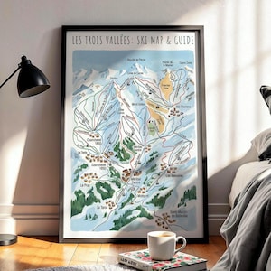 Op de afbeelding: Ingelijste ski-kaart en gids voor Les Trois Vallées, met een gedetailleerde illustratie van skipistes, liften en bergterrein. De kaart staat in een zwart frame, tegen een muur. Een zwarte lamp en een boek met een kopje koffie staan op de voorgrond.