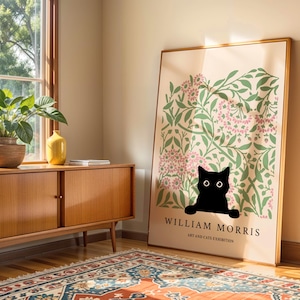 May include: A framed art print featuring a black cat peeking out from a floral design with pink flowers and green leaves. The print has the text "WILLIAM MORRIS ART AND CATS EXHIBITION" in black. The print is leaning against a wall.