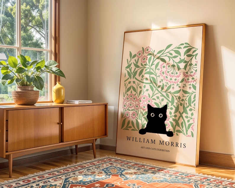 May include: A framed art print featuring a black cat peeking out from a floral design with pink flowers and green leaves. The print has the text "WILLIAM MORRIS ART AND CATS EXHIBITION" in black. The print is leaning against a wall.