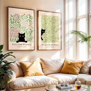 May include: Two framed art prints featuring black cat illustrations. One print has a pink floral background, the other a green leafy design. Both prints have the text "WILLIAM MORRIS" and "ART AND LIFE EXHIBITIONS" in a serif font.