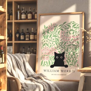 May include: A framed art print featuring a black cat illustration with a floral background in green and pink. The print includes the text "WILLIAM MORRIS ART AND CATS EXHIBITION" in a minimalist style. The artwork is displayed in a room with a wooden shelf.