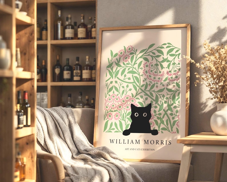 May include: A framed art print featuring a black cat illustration with a floral background in green and pink. The print includes the text "WILLIAM MORRIS ART AND CATS EXHIBITION" in a minimalist style. The artwork is displayed in a room with a wooden shelf.