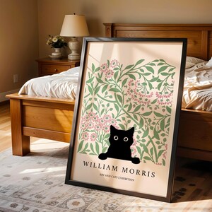 May include: Framed art print featuring a black cat peeking out from behind a floral design in green and pink. The print has the text "WILLIAM MORRIS ART AND CATS EXHIBITION" in black, set against a cream background.