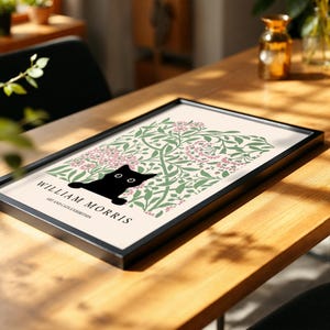 May include: A framed art print featuring a black cat silhouette peeking out from a floral design in green and pink. The print is labeled "WILLIAM MORRIS ART AND CATS EXHIBITION" in black text. The frame is black, and the print rests on a wooden table.