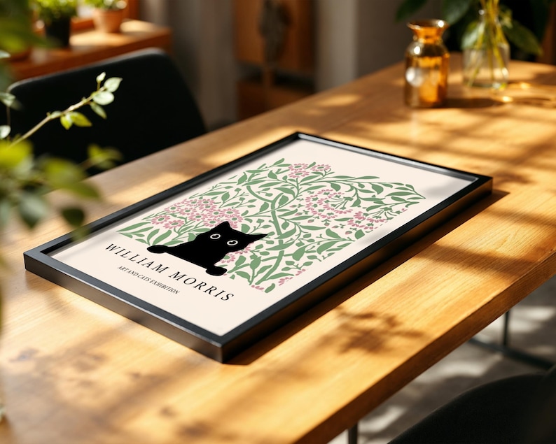 May include: A framed art print featuring a black cat silhouette peeking out from a floral design in green and pink. The print is labeled "WILLIAM MORRIS ART AND CATS EXHIBITION" in black text. The frame is black, and the print rests on a wooden table.