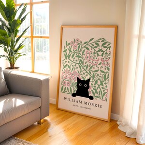 May include: A framed art print featuring a black cat peeking out from a floral design with pink and green accents. The print is labeled "William Morris Art and Cats Exhibition" in a wooden frame, leaning against a wall.
