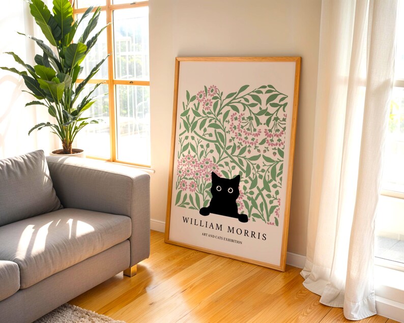 May include: A framed art print featuring a black cat peeking out from a floral design with pink and green accents. The print is labeled "William Morris Art and Cats Exhibition" in a wooden frame, leaning against a wall.
