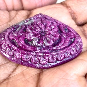 Puede incluir: Una gema tallada en forma de corazón, en tonos magenta oscuro y púrpura. La piedra presenta un diseño floral central rodeado de intrincadas y detalladas tallas. La gema se sostiene en una mano, mostrando su superficie pulida y su artesanía única.