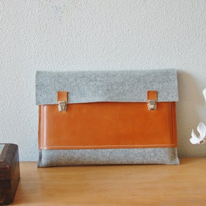 Könnte beinhalten: Eine graue Filz-Laptoptasche mit einer braunen Lederklappe und silbernen Verschlüssen.