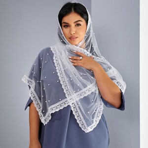 A woman wearing a blue dress with a white lace veil stands against a gray wall, looking directly at the camera with a neutral expression.