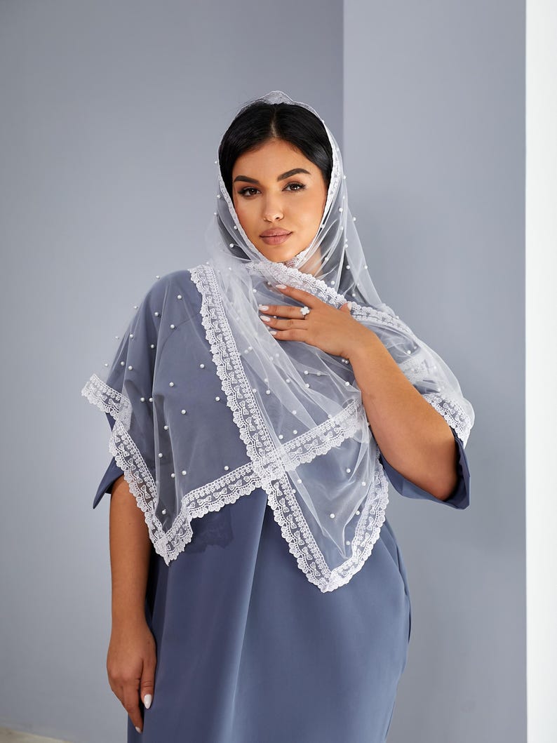 A woman wearing a blue dress with a white lace veil stands against a gray wall, looking directly at the camera with a neutral expression.